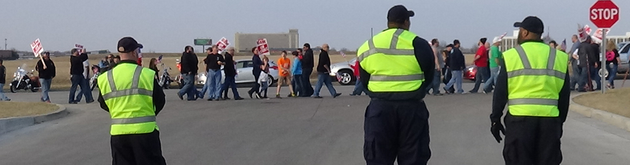 Strike security officers standing at entrance of plant with picketing union members on strike